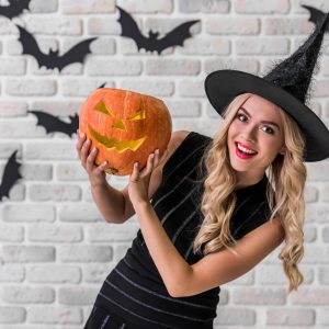 Beautiful blonde girl in black dress and witch hat is holding a scary pumpkin, looking at camera and smiling, on background decorated for Halloween