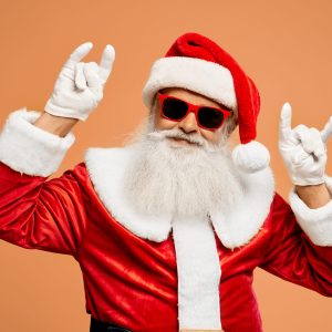 Confident aged bearded Santa Claus in christmas costume and sunglasses looking at camera and showing double horn sign with hands in white gloves. Rock and heavy metal gesture.