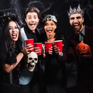 excited multiethnic people with plastic cups laughing at camera during halloween party on black