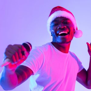 Christmas music. Cool black guy in Santa hat singing holiday song, using microphone, performing karaoke in neon light. Carefree African American singer giving live music concert on New Years eve